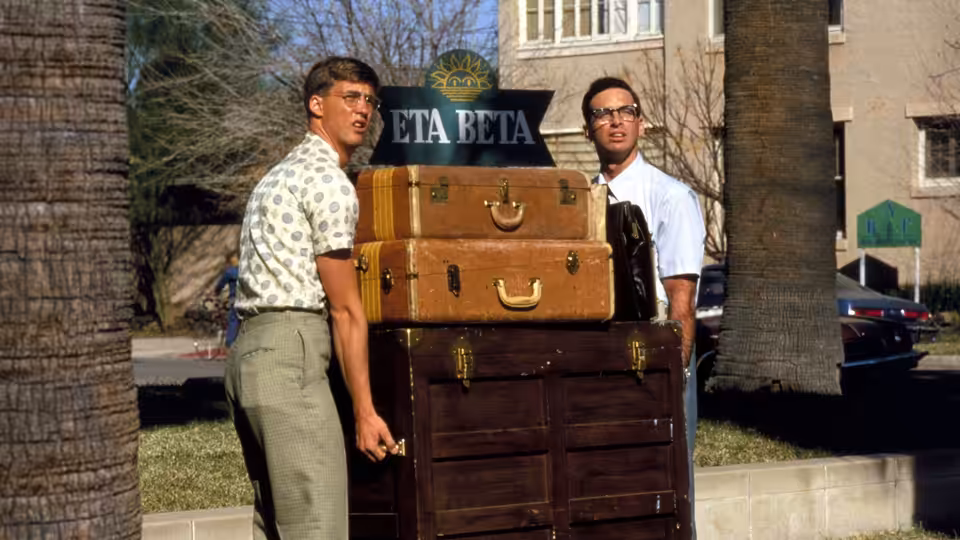 Gilbert Lowell (Anthony Edwards) and Lewis Skolnick (Robert Carradine) carry luggage outside the Eta Beta house