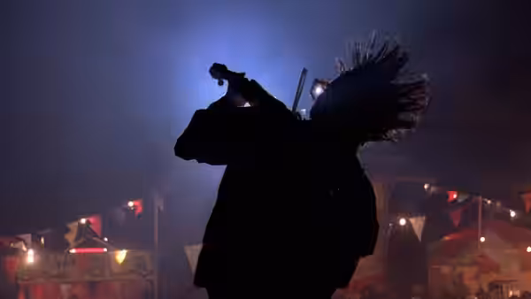 A costumed figure poses in silhouette beneath carnival lights, with booths in the background