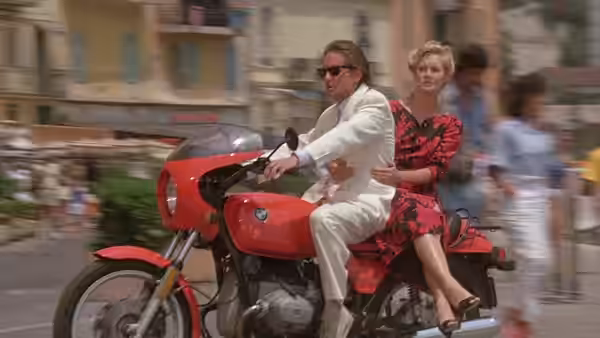 Jack Colton drives a red motorcycle through a crowded street with Joan Wilder riding behind him