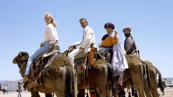 Joan Wilder and Jack Colton ride camels alongside two local guides across open terrain