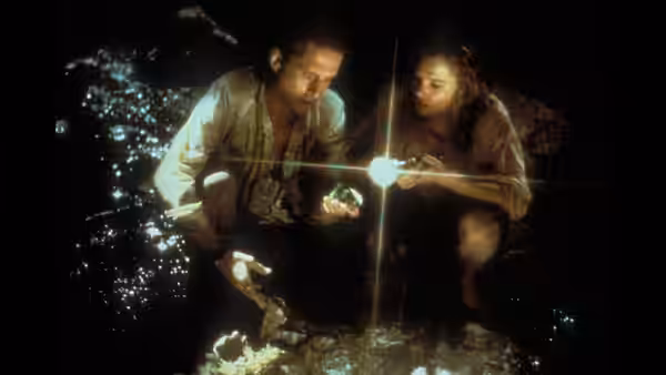Jack Colton and Joan Wilder crouch beside a glowing light over water