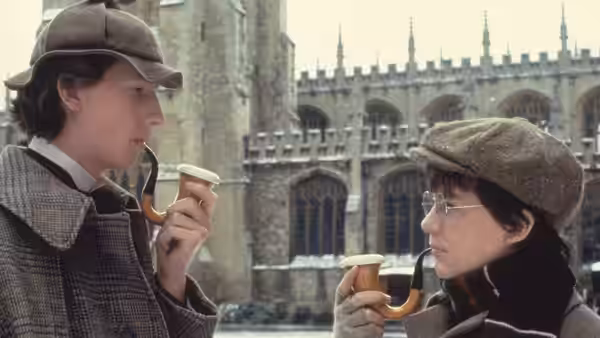 Sherlock Holmes (Nicholas Rowe) and John Watson (Alan Cox) hold pipes outside a Gothic college