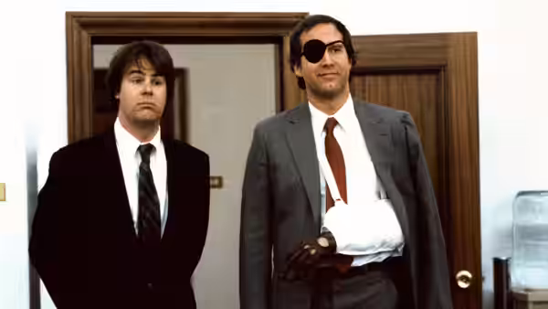 Austin Millbarge (Dan Aykroyd), wearing an eyepatch and sling, stands beside Emmett Fitz-Hume (Chevy Chase) indoors
