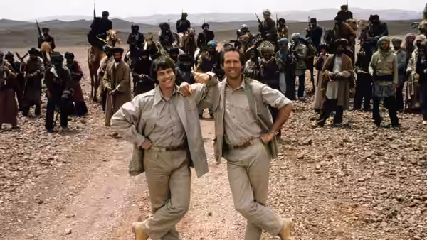 Emmett Fitz-Hume (Chevy Chase) and Austin Millbarge (Dan Aykroyd) pose among armed men in a rocky desert camp