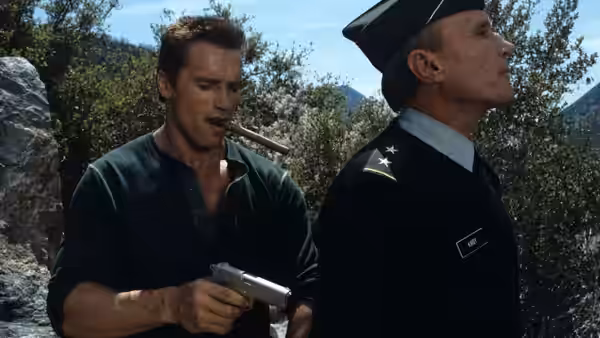 John Matrix (Arnold Schwarzenegger) holds a pistol behind a uniformed officer outdoors