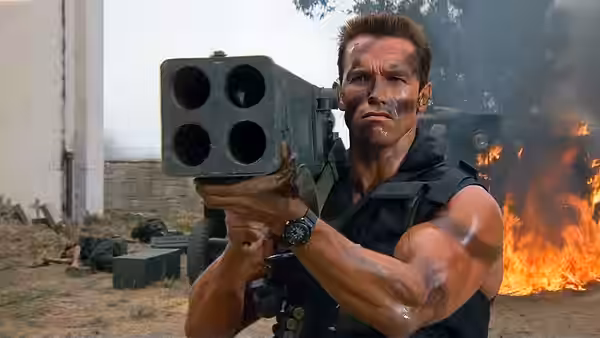 John Matrix (Arnold Schwarzenegger) aims a rocket launcher beside burning wreckage