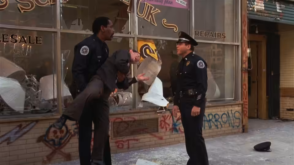 Two uniformed officers examine a vandalized storefront as debris covers the sidewalk