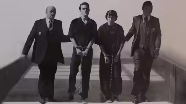 Two handcuffed young men walk between suited escorts along a walkway