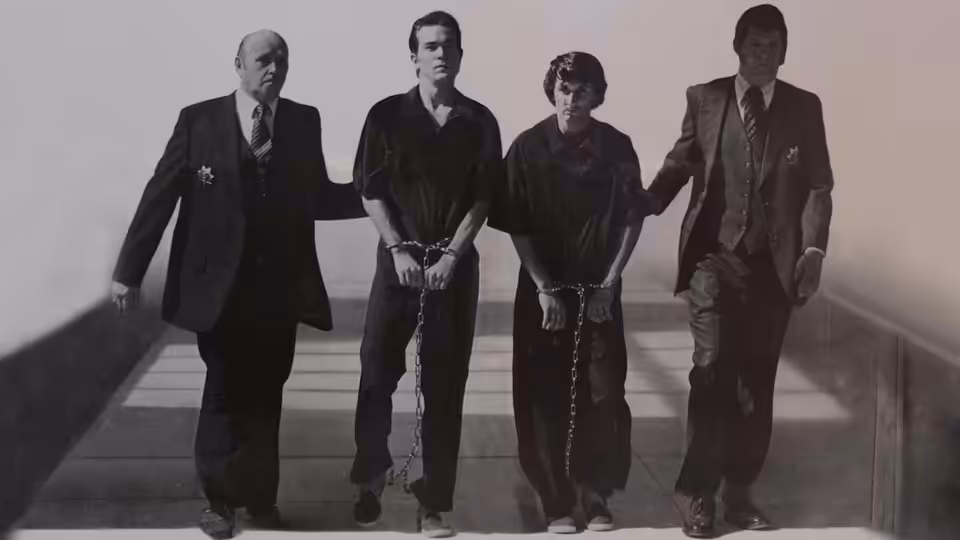 Two handcuffed young men walk between suited escorts along a walkway