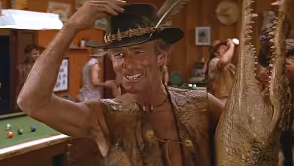 Mick Dundee (Paul Hogan) tips his hat while holding a crocodile in a pub