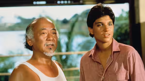 Miyagi (Pat Morita) and Daniel (Ralph Macchio) stand together indoors