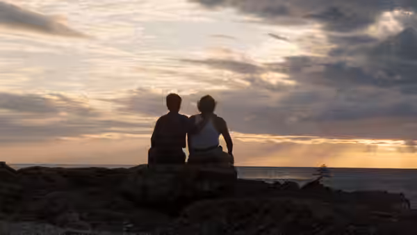 Two people sit together on a rocky shore, watching the sunset
