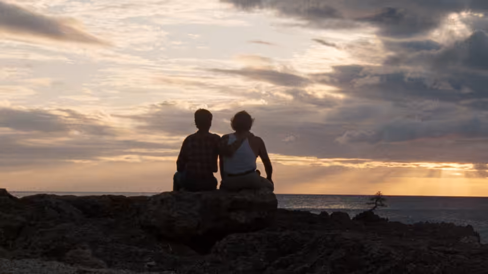 Two people sit together on a rocky shore, watching the sunset
