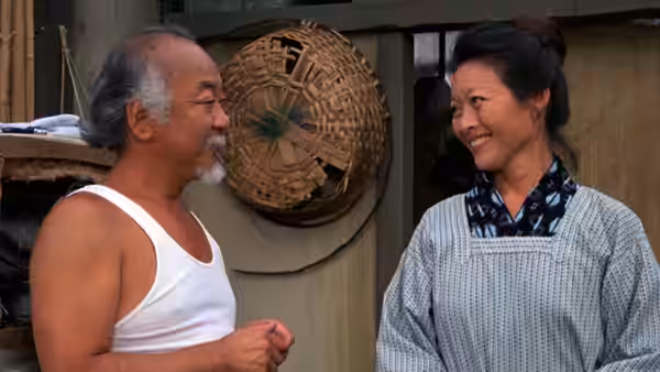 Miyagi (Pat Morita) talks with Yukie (Nobu McCarthy) outside a wooden building