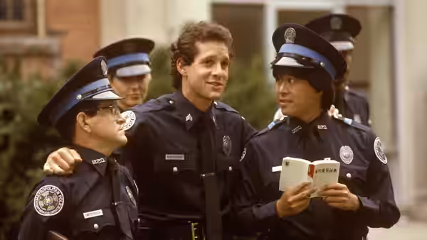 Carey Mahoney (Steve Guttenberg) puts an arm around a bespectacled cadet as Jones reads a book