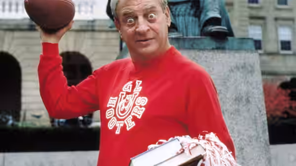 Thornton Melon (Rodney Dangerfield) holds a football and cheerleading pompoms outside