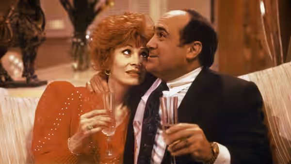 Sam Stone (Danny DeVito) embraces Carol Dodsworth (Anita Morris) as they hold drinks