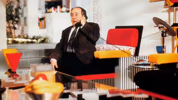 Sam Stone (Danny DeVito) talks on a phone from his colorful office