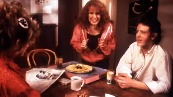 A red-haired woman stands beside a table as a man and another woman sit nearby