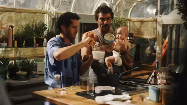 Michael prepares food at a kitchen counter as Peter holds the baby and watches