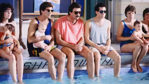 Three men sit by a swimming pool with babies on their laps, while two women sit nearby