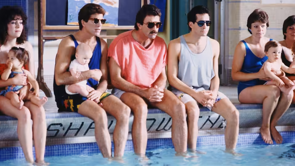 Three men sit by a swimming pool with babies on their laps, while two women sit nearby