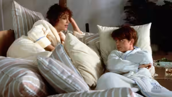 Vera Prescott and Brantley Foster sit wrapped in robes among pillows on a bed