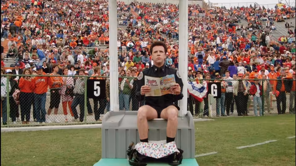 Mahoney (Steve Guttenberg) reads a newspaper while seated on a portable toilet at a stadium