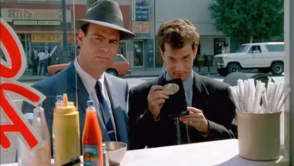 Sgt. Joe Friday (Dan Aykroyd) watches as Pep Streebeck (Tom Hanks) examines a badge at a food stand