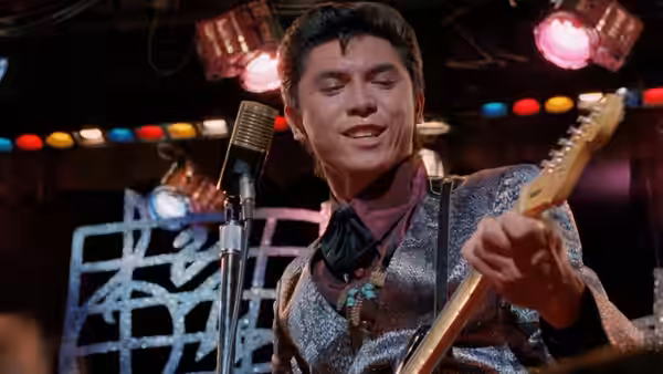 Ritchie Valens (Lou Diamond Phillips) plays guitar and sings into a microphone onstage