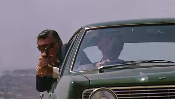 A man aims a handgun beside a green car as another man sits behind the wheel