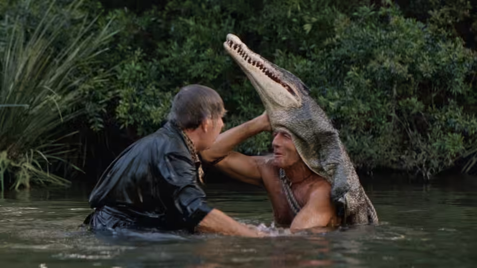 Crocodile Dundee (Paul Hogan) wrestles with a man beside a crocodile in a jungle river