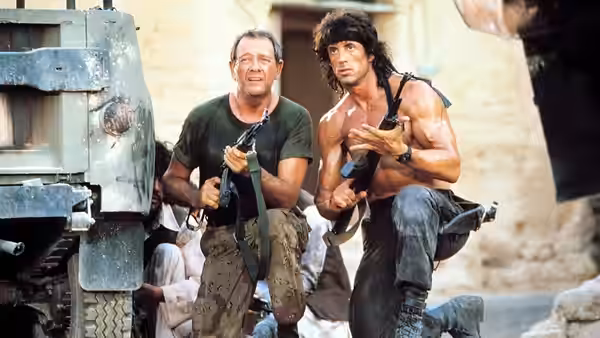 John Rambo (Sylvester Stallone) and Trautman (Richard Crenna) crouch with rifles beside a vehicle