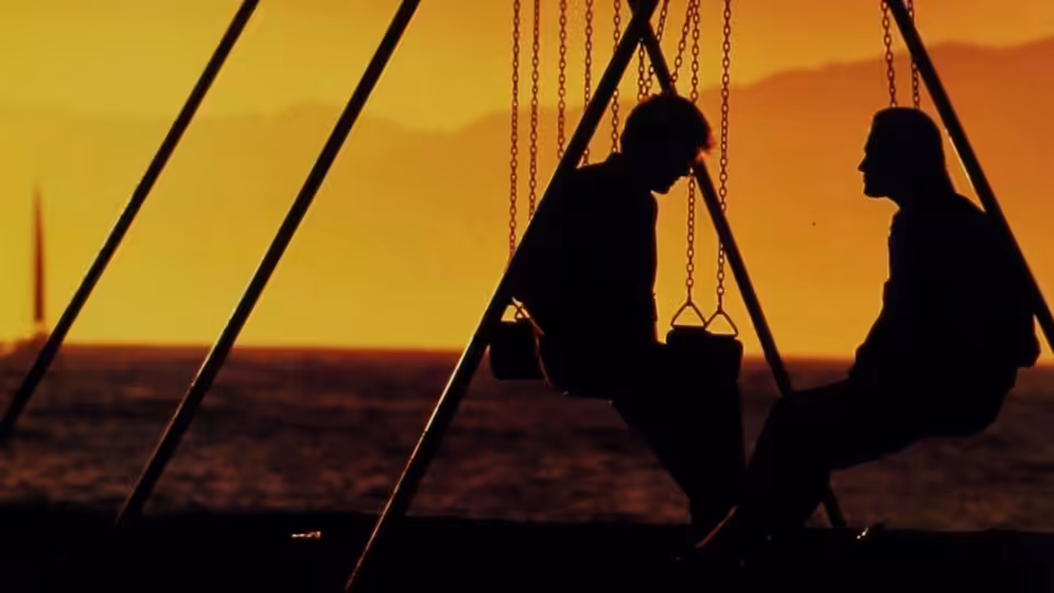 Two silhouetted figures sit facing each other on a boat at sunset