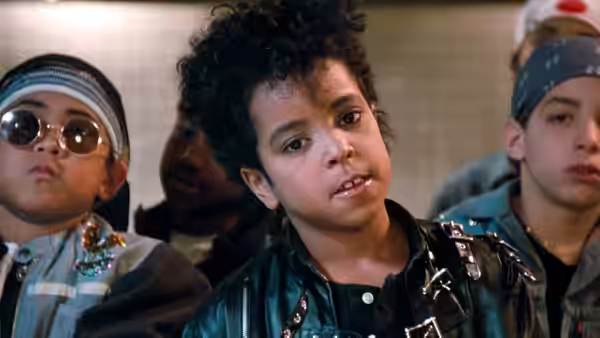 A curly-haired boy in a leather jacket stands among children in bandanas