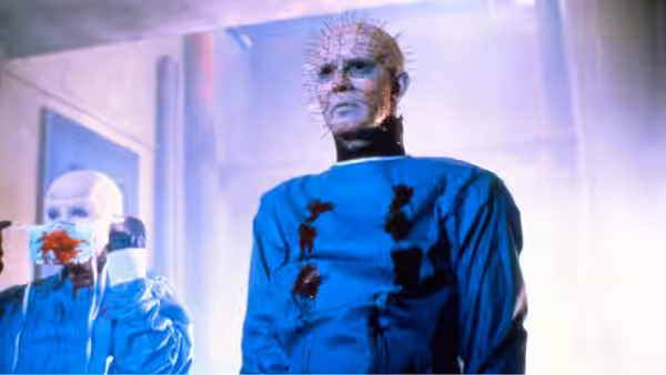 Pinhead (Doug Bradley) stands in a bloodstained blue tunic beside a restrained patient