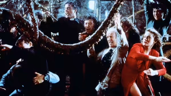 A group of costumed guests recoil and gesture as a huge tentacle stretches across the room