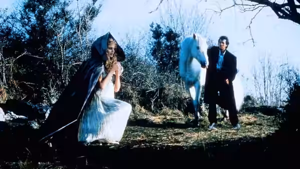 A woman in a white gown and dark cloak crouches beside a man and a white horse