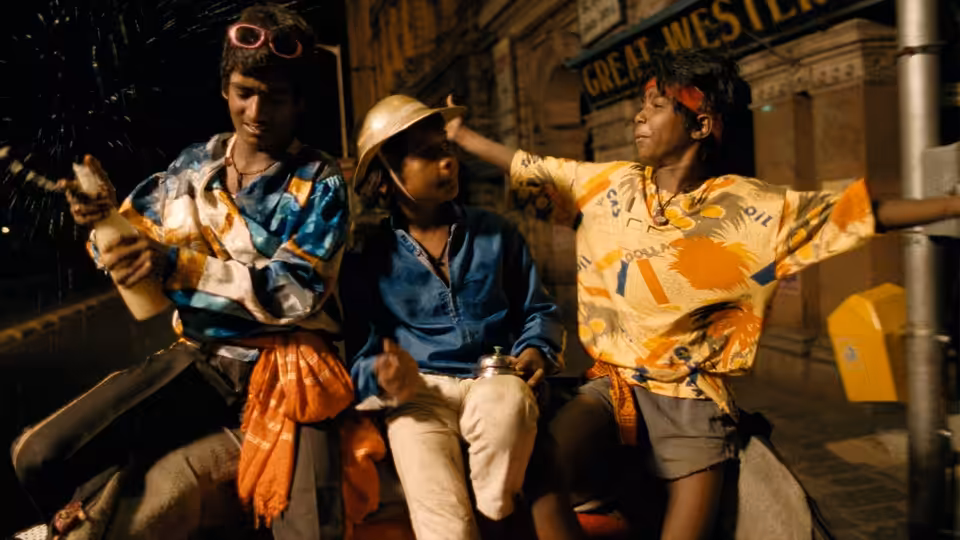 Three boys sit together on a nighttime street, one wearing a helmet