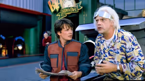 Marty McFly (Michael J. Fox) holds a newspaper beside Doc Brown (Christopher Lloyd) outside a futuristic cafe