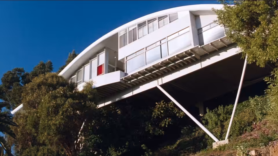 A modern white house stands elevated above trees on tall angled supports