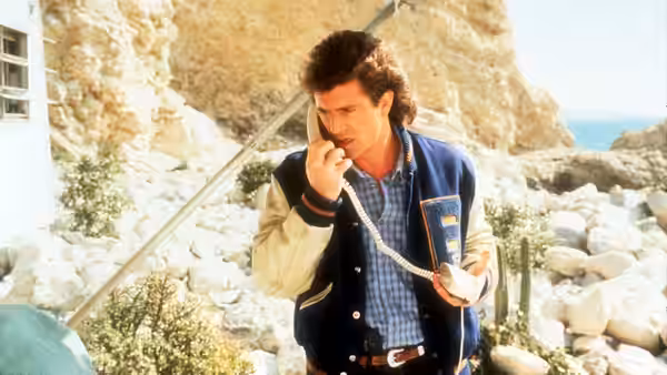Martin Riggs (Mel Gibson) speaks on a telephone outside a rocky coastal house