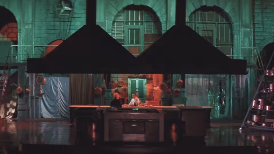 Cooks work behind a broad kitchen counter beneath dark ventilation hoods