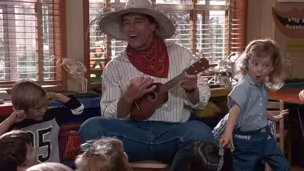 Kimble (Arnold Schwarzenegger) plays ukulele in a cowboy hat as a child dances