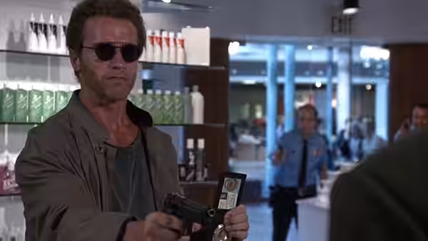 Kimble (Arnold Schwarzenegger), wearing sunglasses, displays his badge and aims a pistol inside a store
