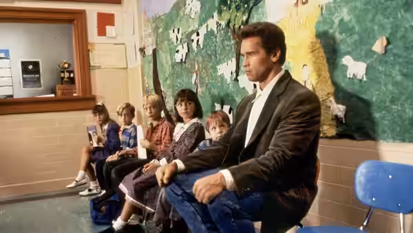 Kimble (Arnold Schwarzenegger) sits beside children waiting along a school hallway