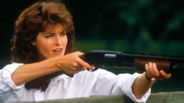 A woman in a white shirt aims a shotgun from behind a wooden barrier