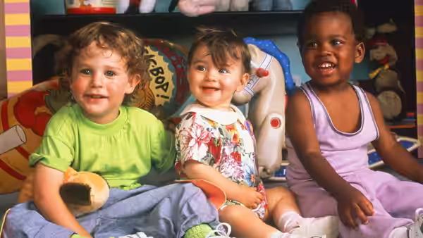 Three young children sit together among toys and cushions