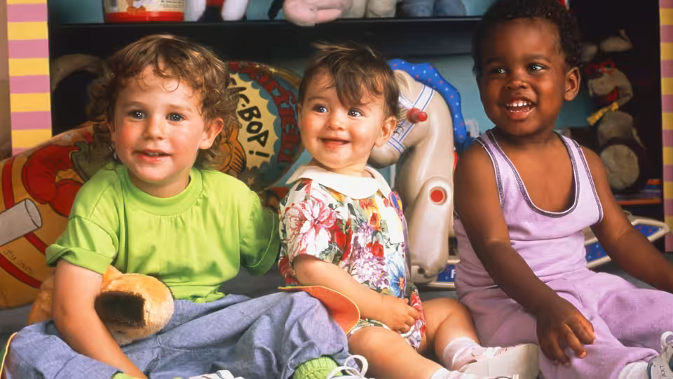 Three young children sit together among toys and cushions