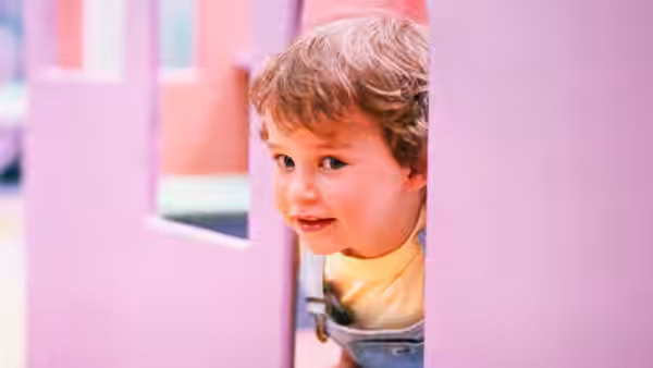 A young child peeks through a narrow gap in a pink door